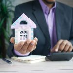 Persona sosteniendo una maqueta de casa junto a una calculadora, haciendo cuentas para decidir si amortizar la hipoteca o invertir sus ahorros